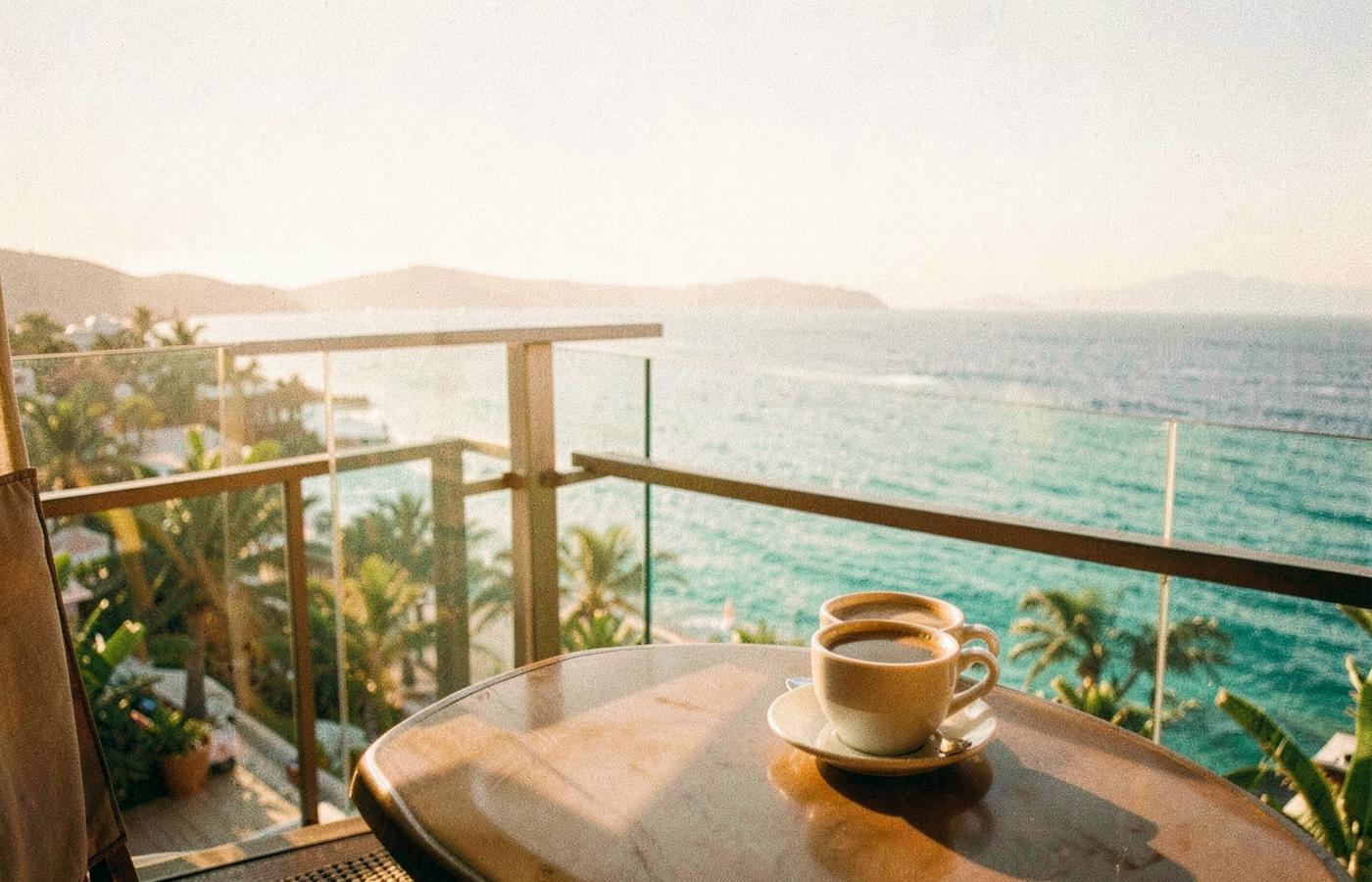 Two coffee cups on a sunlit hotel balcony overlooking a bay